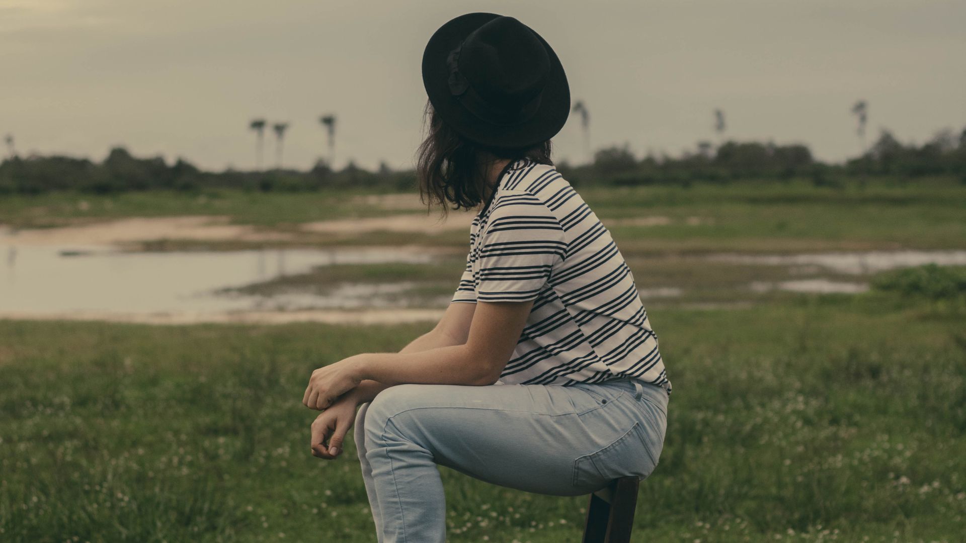 Photo of Woman Sitting on Brown Wooden Chair In the Middle of a Grass Field Looking Away